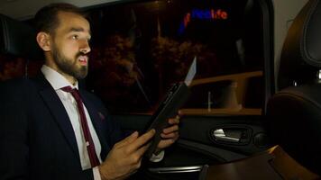 Businessman during a business video call in the back seat of his luxury car at night. Driving through city. Night city lights.