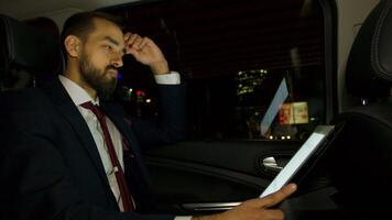 affärsman bär en företag kostym i de tillbaka sittplats av hans lyx bil har en företag video ringa upp. personlig förare. natt stad lampor.