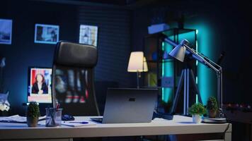 Smart TV left open on news broadcast in empty home office with desk filled with business items. Television set showing VOD channel with Laptop, headphones, plants in blurry foreground, camera B video