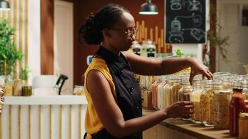 Local merchant organizing homemade pasta in reusable jars at zero waste supermarket, wearing apron and welcoming customers. African american woman in eco friendly store. Camera A. video