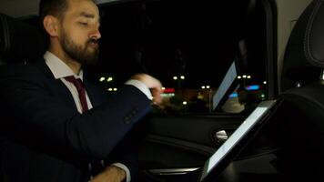 Serious businessman working on tablet at night in the back seat of his limousine with personal driver. Successful businessman. video