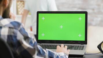 Over the shoulder shot of man working on laptop with green screen display in very bright living space while his roommate sits on the sofa video