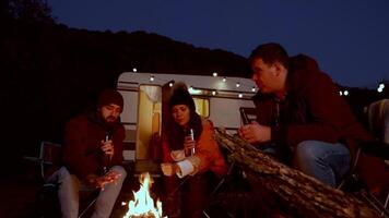 caucásico amigo disfrutando un cerveza juntos en frente de su retro camper y calentamiento su manos a acampar fuego. ligero bombillas video