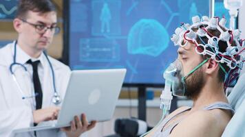 médico utilizando su ordenador portátil en un moderno instalaciones para cerebro investigación. paciente vistiendo aire máscara y ondas cerebrales exploración auriculares. video