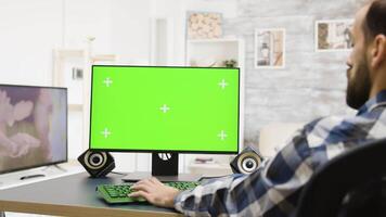 Zoom in shot of man working on green screen PC in bright and well lit apartment while his roommate sits on the sofa watching TV video