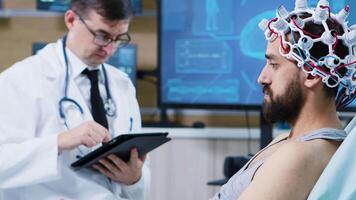 paciente sentado em hospital cama e vestindo ondas cerebrais digitalização fone de ouvido. médico usando tábua. video