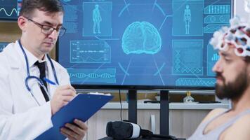 paciente dentro uma moderno clínica para cérebro pesquisar. paciente vestindo ondas cerebrais digitalização fone de ouvido. médico escrevendo em prancheta video