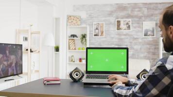 Man browsing the internet on green screen laptop in bright and very well lit flat. In the background - his roommate watches TV video