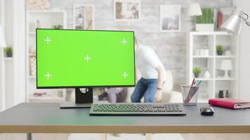 PC with isolated green screen mock-up on the table in bright living room. In the background - two men are watching TV video