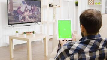 hombre mirando a un película en el televisión mientras participación un verde pantalla digital tableta ordenador personal en su manos. en el antecedentes - brillante y bien iluminado vivo habitación video