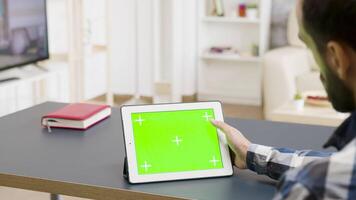 Man scrolling and touching a green screen of a digital tablet in bright living space. His roommate watches a movie on TV in the background video