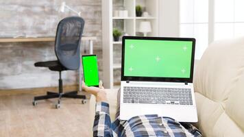 Parallax shot of man lying on the couch, holding a green screen smartphone and laptop in hands in bright and well lit living space video