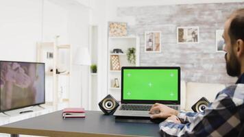 Static shot of man looking at green screen laptop while his roommate watches a movie on the TV video