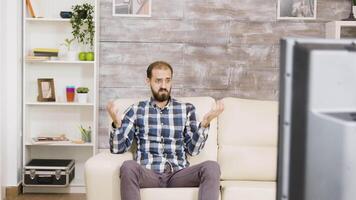 Bearded young man sitting on couch and looking confused at tv. Man in a cozy living room. video