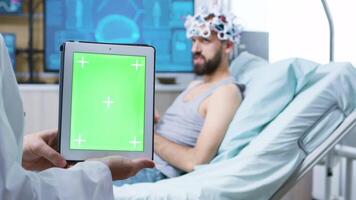 medico nel un' neurologia clinica Tenere digitale tavoletta con verde schermo. paziente indossare onde cerebrali lettura cuffia. video