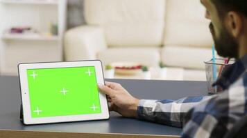 Over the shoulder shot of man tapping on tablet PC with green screen on it in bright and well lit living space video