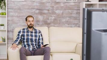 Bearded man sitting with serious face watching tv and holding remote control. Zoom in effect video