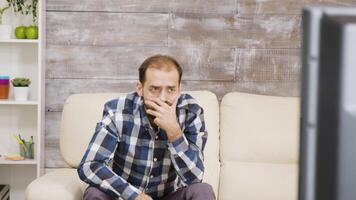 bärtig Mann Sitzung auf Sofa mit schockiert Ausdruck während Aufpassen Fernseher im seine Wohnung. video