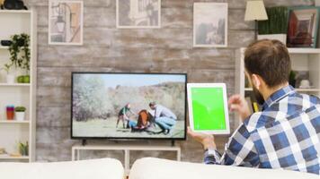 Back view of bearded man sitting on couch holding tablet with green screen. Man swiping on tablet. video