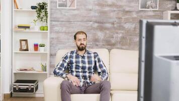Bearded man with curious expression while watching tv sitting on sofa in his apartment. video