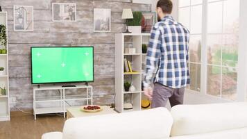 Young man arriving home from work wearing a shirt and watching a movie on green screen TV while sitting on sofa. video