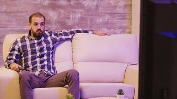 Caucasian bearded man sitting on sofa while watching tv shows. Man relaxing on tv after a long day at work video