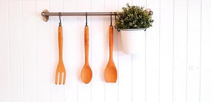 Brown wooden fork, spoon, turner or flipper and green plant in white stainless pot hanging and isolated on white wooden background with copy space. Kitchen utensil, Plant and Vintage object concept photo