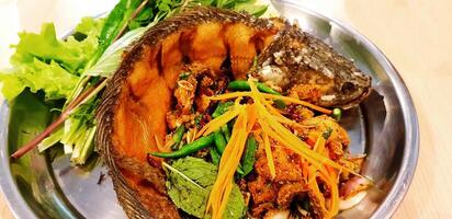 Fried snakehead fish in spicy herb salad with fresh vegetables on stainless steel plate or dish. Asian or Thai food. Selective focus photo