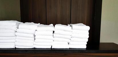 Stack of many clean white towel putting on the black shelf with wooden wall background for customer and tourist take it to shower in the bath room. photo