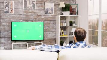 Back view of young man sitting on couch and relaxing while watching a movie on a TV with green screen video