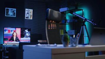 Dimly lit apartment with office desk in front of smart TV displaying news program broadcasting. Laptop on table and television screen in empty room showing channel, camera B close up shot video