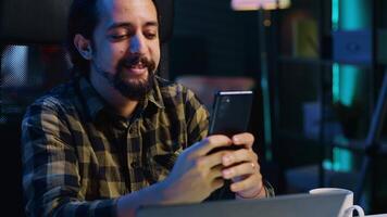 sorridente homem às casa mensagens de texto melhor amigo em Smartphone, levando pausa enquanto trabalhando dentro pessoal escritório. alegre trabalhador autonomo conversando com companheiro conectados usando Móvel telefone, Câmera uma video