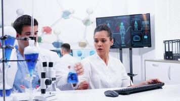 Female scientist in white coat and her assistant working with in modern lab research. Assistant helping female scientist with research. Smoking blue liquid. video