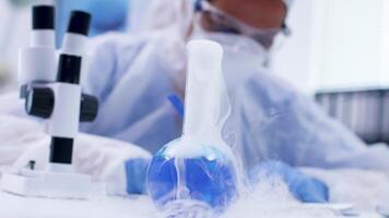 Handheld footage of smoking blue fluid win reagent and scientist looking through microscope. Scientist in white coverall equipment. video