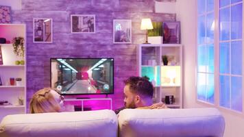 Couple enjoying their time together and playing online video games sitting on couch using wireless controllers.