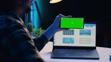 Man sitting on chair at home office desk in neon illuminated apartment holding green screen phone while working. Person in living room looking at business graphs on laptop and at mockup smartphone video