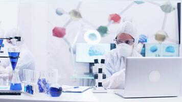Woman scientist looking through microscope in laboratory wearing protection equipment. Scientist taking a flask with reactive blue solution. video
