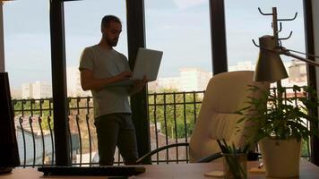 Employee of start up company working on the laptop near big windows in the office. Warm sunset light hitting from behind. Handheld slow motion shot video