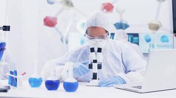 Female scientist working in a modern laboratory with reagent in test tubes with blue liquid and adjusting microscope. Scientist using laptop for research. video