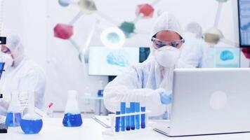 Female scientist picking up a test tube with blue solution and typing on computer. Scientist in protection uniform. video