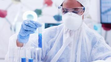 Female researcher in coverall equipment holding a test tube with smoking blue solution. Scientist typing on computer. video