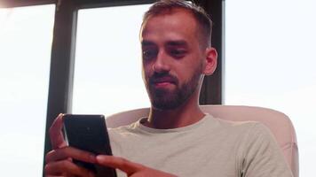 Close up shot of young man smiling while looking at the smartphone screen near a big window. Warm sunset light. Slow motion shot video