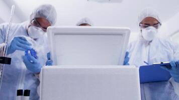 Team of researcher in a modern laboratory opening refrigerator box and taking notes. Scientist in protection equipment. video