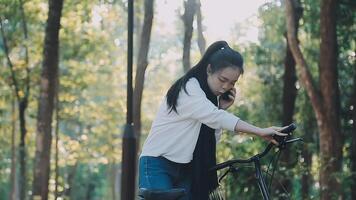 gelukkig vrouw in jeans kleren staan in de buurt fiets fiets Aan trottoir in groen park buitenshuis gebruik mobiel cel telefoon 13 pro babbelen online. mensen actief stedelijk gezond levensstijl wielersport concept video