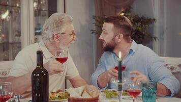 Bij een familie diner, twee mannen drinken wijn en lachen. vader en opa praten en grap Bij een familie viering. geroosterd brood. gelukkig familie. de verhaal van een groot familie. video