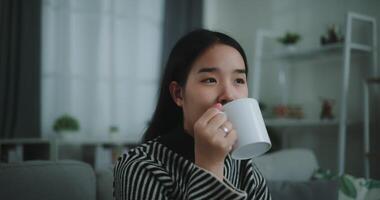 metraggio costante tiro, selettivo messa a fuoco di rilassante giovane donna Tenere tazza godere odore caffè o tè e potabile con felicità nel mattina mentre sedersi su divano nel vivente stanza, libera tempo, prendi rompere nel casa video