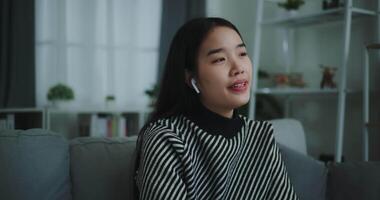 métrage de face vue de content Jeune asiatique femme prendre plaisir écoute à le la musique avec sans fil écouteurs tandis que séance boisson café sur canapé dans vivant pièce à maison, loisir et style de vie, gratuit temps video