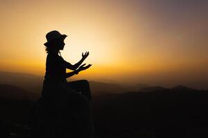 silhuette mujer joven rezando en la montaña, con los brazos extendidos observando un hermoso amanecer dramático. foto