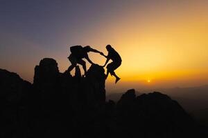 siluetas de dos personas alpinismo en montaña y Ayudar. trabajo en equipo de dos hombres caminante Ayudar cada otro en parte superior de montaña alpinismo equipo hermosa amanecer. ayuda y asistencia concepto. foto