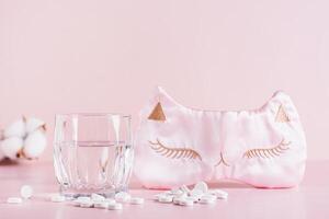 Sleeping pills, a glass of water and a sleep mask on a pink background photo